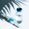 small glass ampoule, clear liquid, blue syringe resting on top, bright white background, shadow of a fern in background